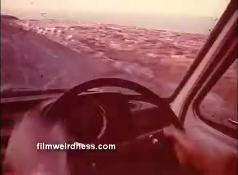 The still depicts a close-up view of hands gripping a steering wheel, suggesting a dynamic driving scene. The landscape outside is hazy and blurred, indicating speed or motion, adding a sense of urgency. The warm color tones contribute to a vintage feel typical of 1970s cinema. This moment likely evokes suspense or excitement, characteristic of a thriller backdrop.