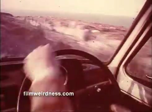 The still from the trailer of 'The Mackintosh Man' features a close-up of a driver's hand gripping the steering wheel of a vehicle, suggesting a sense of urgency or tension. The blurred landscape outside indicates movement, likely conveying a journey or chase. The warm, faded colors evoke a vintage aesthetic typical of 16mm film, enhancing the dramatic tone associated with the cinematic work starring Paul Newman.