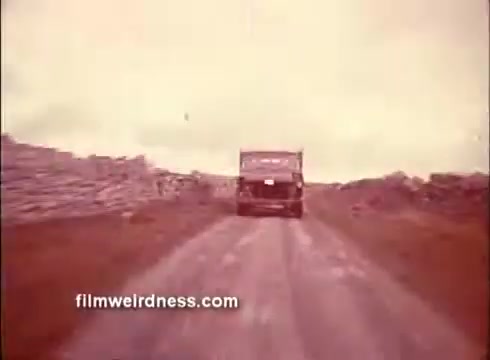 The still from 'The Mackintosh Man' trailer shows a truck driving down a dirt road, flanked by stone walls or barriers. The scene is set against a cloudy sky, giving it an atmospheric and somewhat tense feel, typical of suspenseful storytelling. The focus is on the vehicle's journey, hinting at themes of pursuit or escape within the film.