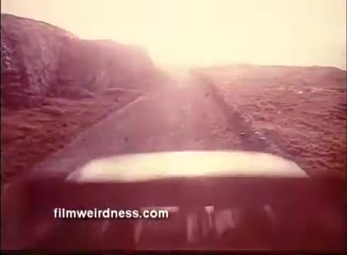 The still from 'The Mackintosh Man' trailer features a sun-drenched gravel road, suggesting a sense of journey or adventure. The view appears to be from a vehicle, emphasizing the isolation of the landscape, with rocky terrain flanking the pathway. The light gives the scene a dreamy, almost surreal quality, hinting at the film's themes of suspense and intrigue.