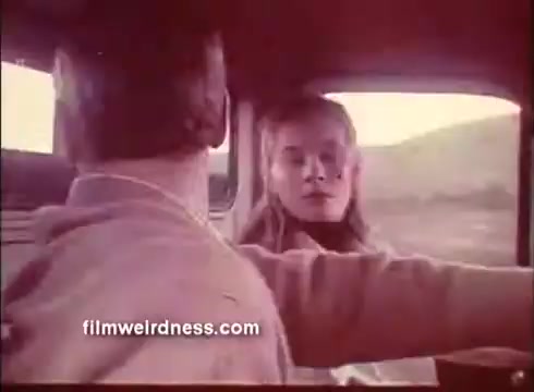 The still from 'The Mackintosh Man' trailer features two characters inside a vehicle. The focus is on a woman with long hair, looking intently at the male driver, who is partially visible. The setting appears to be rural, with a blurred landscape outside the windows, suggesting a moment of tension or conversation between them. The color palette has a vintage feel, typical of the film's era.