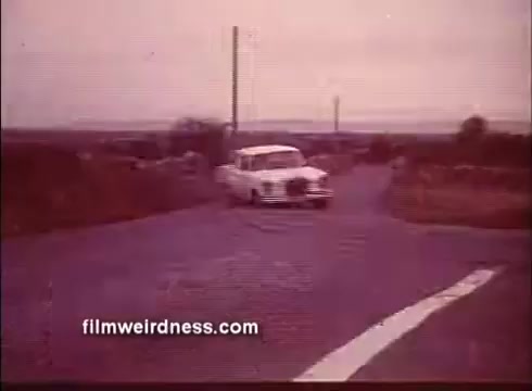 The still from the trailer of 'The Mackintosh Man' features a vintage car turning onto a rural road, set against a soft, muted background. The landscape appears desolate, with sparse vegetation and distant hills, contributing to a sense of isolation. The overall color palette is warm, evoking a nostalgic atmosphere typical of films from the era.