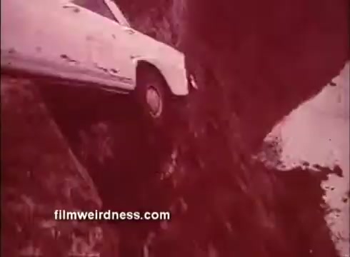 The still from 'The Mackintosh Man' trailer features a white car precariously perched on the edge of a rocky cliff. The scene is tinted in a reddish hue, adding to the intensity and tension of the situation. This moment likely captures a dramatic or suspenseful moment from the film, highlighting themes of danger and high stakes.