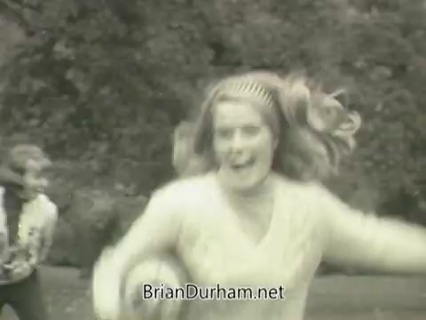 The still features a young woman with long, flowing hair and a bright expression, playing in an outdoor setting, likely a park. She is wearing a light-colored sweater and appears to be running or engaged in playful activity, with a male figure partly visible in the background. The overall tone is lively and carefree, capturing a moment of fun and youthfulness, typical of advertising in that era.