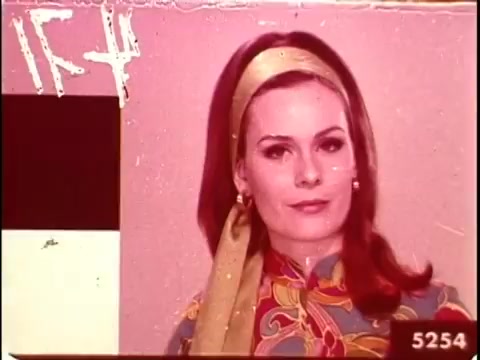The still features a woman in vibrant, patterned attire typical of mid-20th century fashion, with a notable headscarf. This image is part of a 16mm film leader, used historically to assess color accuracy in film development. The background is a soft pink, with a color test card on the left. The frame is marked with numbers, indicating it's part of a sequence for evaluation purposes.