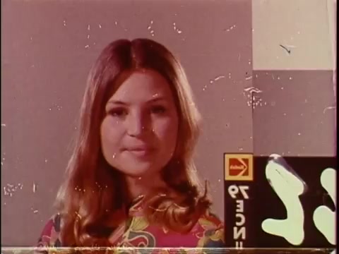 The still features a young woman known as a 'China girl,' a term used in film to denote test patterns for color correction in 16mm film stock. She has long, wavy hair and is dressed in a vibrant, patterned outfit typical of the era. The background includes a color calibration element, often used in film processing, along with a film lab logo. The image shows the characteristic texture of aged or vintage film, with scratches and marks present.