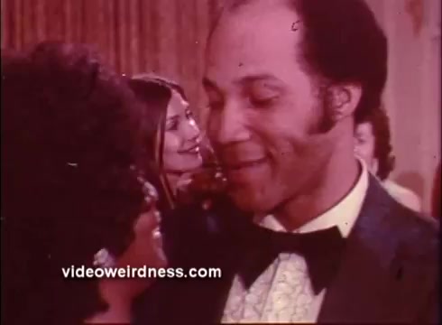 The still from 'Come Back, Charleston Blue' features two characters in close conversation, likely at a social event. The foreground shows a man in a bowtie smiling warmly at a woman, while behind them, another woman appears to be engaged in the moment as well. The scene suggests a lively, intimate atmosphere typical of the blaxploitation genre, emphasizing personal connections amid a vibrant social backdrop. The 16mm print quality adds a vintage feel, characteristic of the era's filmmaking style.