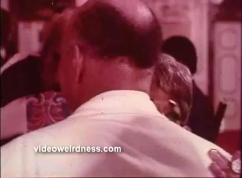 The still from 'Come Back, Charleston Blue' is a close-up of a man's back, highlighting his bald head and the collar of his shirt. In the background, figures are partially visible, suggesting a busy, social atmosphere typical of Blaxploitation films. The colorful patterns of clothing hint at the vibrant style of the era. The image captures a moment of interaction, reinforcing the communal and lively themes prevalent in the genre.