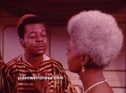 The still shows a scene from the Blaxploitation film 'Come Back, Charleston Blue.' Two characters are depicted in a close-up interaction; one man wears a patterned garment and has a contemplative expression, while the woman, adorned with a pearl necklace, sports a strikingly coiffed gray afro. The setting has a vibrant, colorful backdrop that reflects the film's distinctive aesthetic. The image captures a moment of emotional exchange between the characters.