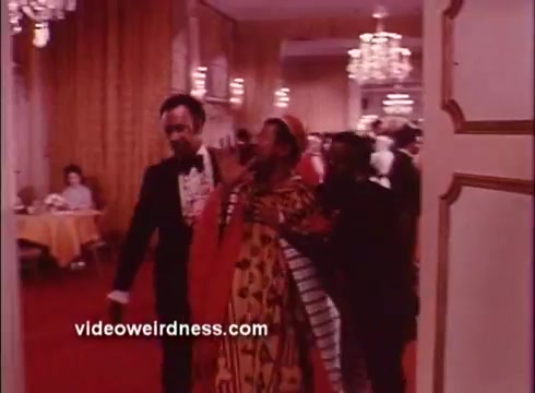 The still from 'Come Back, Charleston Blue' features a vibrant indoor scene, likely in a social or dining setting. A man in a tuxedo leads another man dressed in a colorful, patterned outfit, suggestive of traditional or cultural attire. The background reveals a banquet of elegantly set tables and chandeliers, contributing to the film's retro ambience. Overall, the scene embodies the theatrical and stylish essence typical of Blaxploitation films.