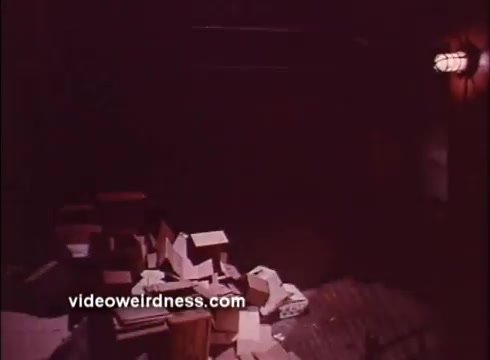 The still from 'Come Back, Charleston Blue' features a dimly lit scene with disordered piles of boxes and debris scattered on the ground. The lighting is moody, enhancing the film's gritty atmosphere. The overall tone suggests a setting that is potentially abandoned or neglected, characteristic of the blaxploitation genre's urban themes.