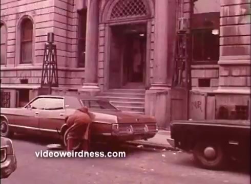 The still from 'Come Back, Charleston Blue' features a scene set on a city street, showcasing a vintage automobile parked prominently. A person, dressed in a brown coat, is seen bending down near the back of the car, possibly interacting with it. In the background, a building with arched windows and decorative architecture is visible, adding to the urban atmosphere. The overall color palette is muted, characteristic of the 16mm film format, conveying a gritty, nostalgic vibe typical of the Blaxploitation genre.