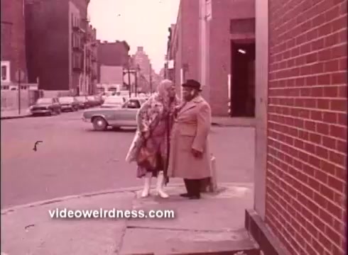 The still from 'Come Back, Charleston Blue' features two characters conversing at a street corner. The woman, dressed in a vibrant outfit with a fur coat and white boots, stands close to a man in a large overcoat and hat. The background shows a cityscape with parked cars and buildings, capturing a vintage urban ambiance typical of the Blaxploitation era. The color tone is muted, giving a nostalgic feel to the scene.