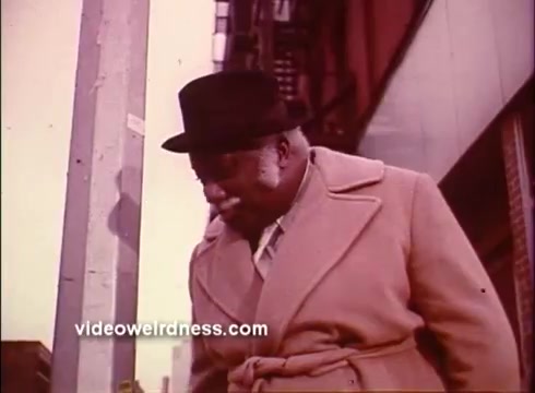 The still from 'Come Back, Charleston Blue' features a distinguished elderly man dressed in a light-colored coat and a black fedora. He is walking down a city street, seemingly deep in thought, with a focus on his contemplative expression. The urban backdrop suggests a lively setting typical of the Blaxploitation genre. The color palette is warm, characteristic of the era's film style, emphasizing a nostalgic vibe.