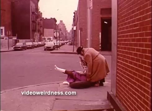 The still from *Come Back, Charleston Blue* features a street scene where two women are positioned on the ground. One woman appears to be assisting the other, who is lying down, dressed in a purple outfit. The background shows a city street with parked cars and buildings, indicated by a warm, vintage color palette typical of 16mm film. This promotional shot encapsulates a moment of drama, characteristic of the Blaxploitation genre.