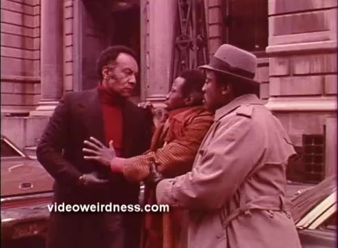 The still from *Come Back, Charleston Blue* features three male characters engaged in a tense interaction on a city street. One man, wearing a black coat and red turtleneck, appears to confront another, who is dressed in a brown coat and is being held back by a third character in a trench coat and hat. The backdrop includes urban architecture, contributing to the film's Blaxploitation aesthetic. The overall atmosphere suggests conflict or drama among the characters.