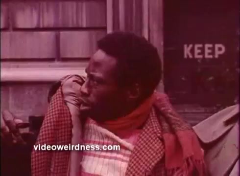 The still from 'Come Back, Charleston Blue' features a man in a patterned scarf and coat, looking off-screen with a curious or intense expression. The setting appears to be an outdoor area, possibly suburban, with a wooden structure in the background. A sign that reads 'KEEP' is visible, adding to the scene's intrigue. The overall tone reflects the stylistic elements of Blaxploitation cinema, emphasizing character and mood. The image is tinted with a vintage, muted color palette typical of 16mm film.