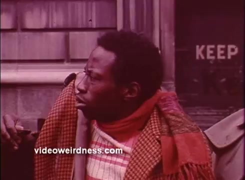 The still from 'Come Back, Charleston Blue' features a man with short hair, wearing a colorful scarf and a patterned jacket. He gazes thoughtfully to the side, surrounded by a rustic urban setting. The background hints at a worn building, with the word 'KEEP' visible, suggesting a moment that reflects both tension and contemplation. The overall tone of the image is warm, emphasizing the character's expression and attire in a vintage style typical of Blaxploitation films.