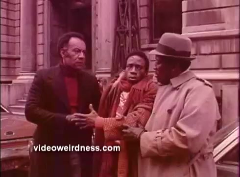 The still depicts three men engaged in a conversation on the street, likely reflecting tension or urgency. One man, dressed in a red sweater under a black jacket, appears stern, while another, wearing an orange scarf, looks distressed or concerned. The third man, in a light coat and hat, appears to be mediating between the first two. The backdrop shows an urban setting with a classic building, hinting at the film's Blaxploitation themes. The color tone suggests an aged 16mm print, characteristic of promotional materials from that era.