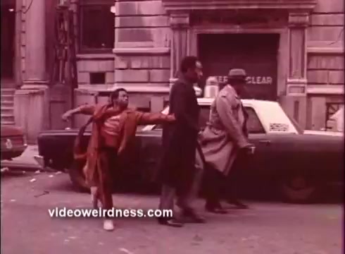 The still from 'Come Back, Charleston Blue' features three men walking along a city street, with a classic car parked nearby. One man, dressed in a vibrant red outfit, appears to be animatedly gesturing, while the others maintain a cooler demeanor in suits and a trench coat. The setting reflects the urban vibe typical of the Blaxploitation genre, and the color palette, likely from the 16mm print, has a warm, vintage tone. The scene suggests a dynamic moment, hinting at themes of camaraderie and street life.