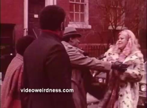 The still from 'Come Back, Charleston Blue' features a group of individuals engaged in a dynamic interaction outside a building. One character, dressed in a trench coat and a hat, appears to be reaching out toward a woman with light, wavy hair adorned in a patterned coat. Another person nearby observes the scene, adding to the sense of tension or drama. The warm tones and vintage look reflect the film's Blaxploitation genre.