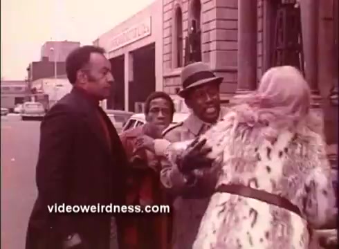 The still from 'Come Back, Charleston Blue' captures a tense street scene featuring three men and a woman. The front characters are engaged in a heated interaction, showing expressions of concern and frustration. The setting appears urban, with vintage cars and buildings in the background, reflecting the film's 1970s context. The woman, dressed in a fur coat, seems to be the focus of the confrontation, while one of the men reaches out toward her. The overall tone suggests themes of conflict and drama typical of Blaxploitation films.