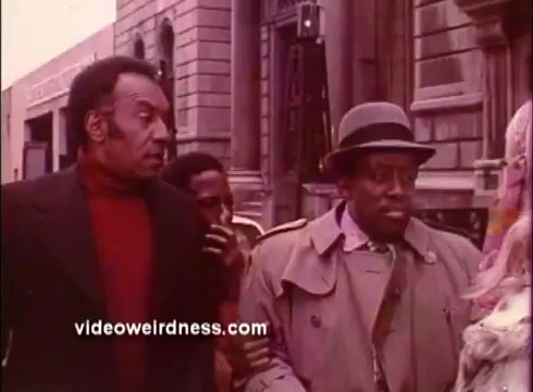 The still features two men walking down a city street, with one character in a red turtleneck and the other in a light trench coat and hat. A third figure is partially visible behind them. The setting exudes a vintage urban atmosphere typical of Blaxploitation films. The scene captures a moment of interaction and conveys a sense of camaraderie or tension amid the urban backdrop. The color saturation reflects the film's 1970s aesthetic.