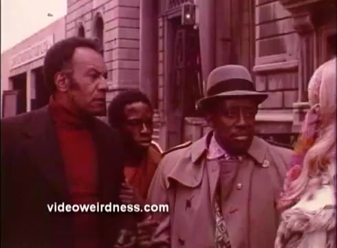 The still from 'Come Back, Charleston Blue' features three central characters engaged in conversation on a street. The first character, wearing a red turtleneck, stands to the left with a serious expression. A younger person peeks from behind him, while the third character, dressed in a light-colored coat and hat, appears to be animatedly speaking. The setting suggests a vibrant urban environment typical of the Blaxploitation genre. The overall tone hints at a blend of drama and community dynamics.