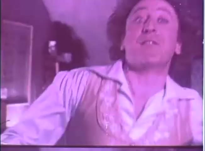 The still from the trailer of 'The Adventure of Sherlock Holmes' Smarter Brother' features a man with an expressive face, caught in a moment of exaggerated emotion. He wears a light-colored vest over a collared shirt and has wild hair, suggesting a comedic or frantic situation. The background has a purple hue, adding a whimsical or surreal atmosphere. Overall, the image conveys a humorous and dynamic scene likely characteristic of the film's comedic tone.