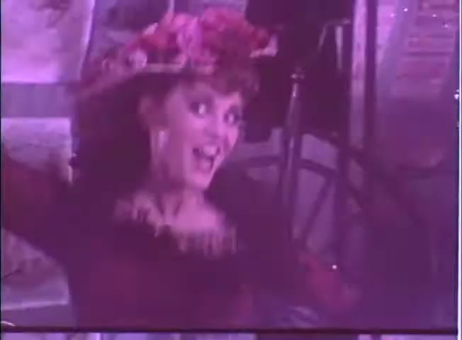 The still features a woman wearing a large, flower-adorned hat, smiling and appearing lively. She embodies a playful, spirited character, reflecting the comedic tone of the film. The visual style has a vintage quality, typical of a 16mm film, contributing to a whimsical atmosphere that aligns with the light-hearted themes of 'The Adventure of Sherlock Holmes' Smarter Brother.'