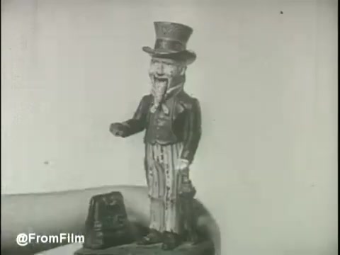 The still features a whimsical, vintage toy bank resembling a character in formal attire, complete with a top hat. It stands on a flat surface, and a hand can be seen nearby, emphasizing its small size. The background is plain, directing attention to the toy, which likely symbolizes saving and financial literacy, aligning with the theme of US Savings Bonds in the 1960s.   Follow us on Twitter @FromFilm.