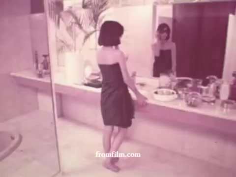 The still is from a 1970s Brazilian Ray-Ban commercial, depicting a woman in a bathroom setting. She wears a simple strapless dress and stands by a sleek, modern sink, gazing at her reflection in a large mirror. The bathroom features a minimalist design with light-colored tiles and a few decorative elements, embodying the aesthetic of the era. The overall tone is soft and nostalgic, highlighting both the fashion of the time and the elegance associated with Ray-Ban sunglasses.