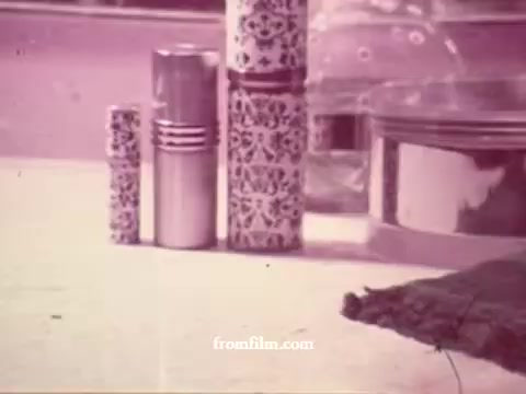 The still features a tabletop arrangement of decorative containers or bottles in a muted color palette typical of the 1970s aesthetic. The foreground showcases various cylindrical objects with intricate designs, reflecting a vintage style. A smooth surface beneath them enhances the overall retro vibe, while a blurred background hints at additional context, possibly related to a lifestyle or fashion theme. The composition suggests a focus on aesthetics, characteristic of commercial advertising from that era.