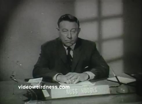 The still features a somber-looking man seated at a desk with a nameplate reading 'RUSS HODGES.' The setting has a muted color palette, indicative of the 1950s era, and includes a desk with scattered papers and a pen. The lighting creates a shadowy ambiance, adding to the serious tone of the public service announcement for the Air Force.