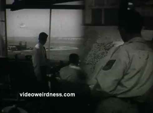 The still depicts a scene from a 1950s Public Service Announcement featuring military personnel in a control room. One officer stands next to a map while two others are seated and focused on monitors, likely overseeing air traffic or operations. The background shows an airfield, emphasizing the military setting and the importance of air defense during that era.