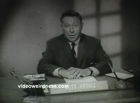 The still features Russ Hodges seated at a desk, likely addressing an audience in a public service announcement for the Air Force from the 1950s. He appears serious and professional, dressed in a suit and tie. The setting is simple, emphasizing the focus on his message, with a nameplate reading 'RUSS HODGES' in front of him. The lighting creates a somber atmosphere, suitable for the context of a PSA.