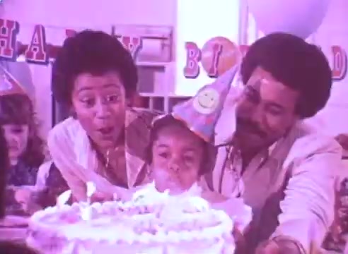 The still depicts a joyful birthday celebration featuring a young child blowing out candles on a cake, surrounded by two adults who appear excited and supportive. The atmosphere is festive, emphasized by party hats and a 'Happy Birthday' banner in the background, creating a warm and happy familial scene that aligns with the theme of celebrating health and well-being in the context of taking medication.