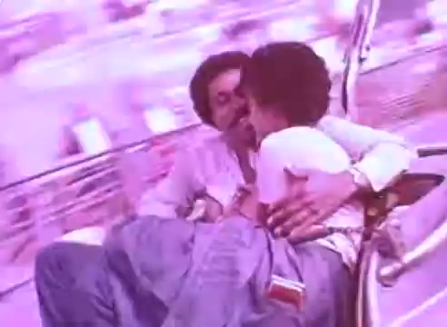 The still depicts two individuals on a ride, engaged in a tender moment. The atmosphere is vibrant, with a pinkish hue giving a dreamy quality to the scene. The context suggests a focus on the importance of health and well-being, framed within a nostalgic, intimate setting. The imagery likely aims to convey the emotional connections that motivate individuals to maintain their health, particularly by taking blood pressure medication.