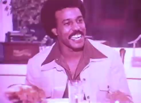 The still features a smiling man with an afro, seated at a dining table. He wears a light-colored jacket over a brown shirt, conveying a cheerful demeanor. The scene is warm and intimate, possibly capturing a moment of family or community gathering. The overall tone suggests a positive message related to health and family, aligned with the campaign's focus on taking blood pressure medication for loved ones.