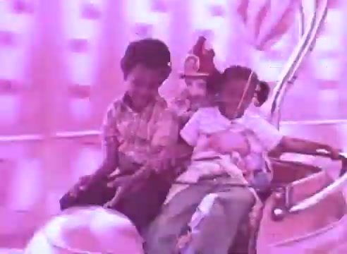 The still features three children enjoying a ride, with a whimsical, colorful background. One child is dressed in a playful outfit, possibly resembling a character or clown. The atmosphere is light-hearted and joyful, emphasizing fun and innocence, aligned with the theme of encouraging health and well-being through medication. The predominant color is a vibrant pink, contributing to a cheerful ambiance.