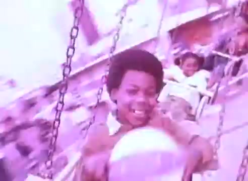 The still from 'Do it for them' - take your blood pressure medication 2 depicts children joyfully riding on swings at a carnival or amusement park. The atmosphere is colorful and lively, capturing moments of happiness and carefree play. The focus is on a young boy with an expressive smile, emphasizing the joy of childhood. Overall, the image illustrates the importance of health and well-being through the lens of joyful experiences.