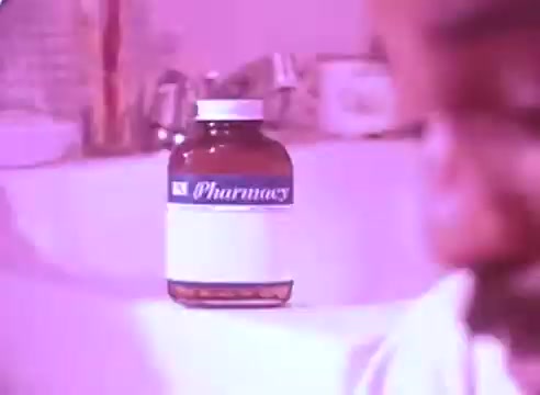 The still features a prescription bottle labeled 'Pharmacy' prominently in the foreground, likely containing blood pressure medication. The background is softly blurred, possibly depicting a bathroom setting. The color filter gives the scene a pinkish hue, creating a calm but serious atmosphere, emphasizing the importance of medication for health. The context suggests a reminder to take medications for better health, particularly for blood pressure management.