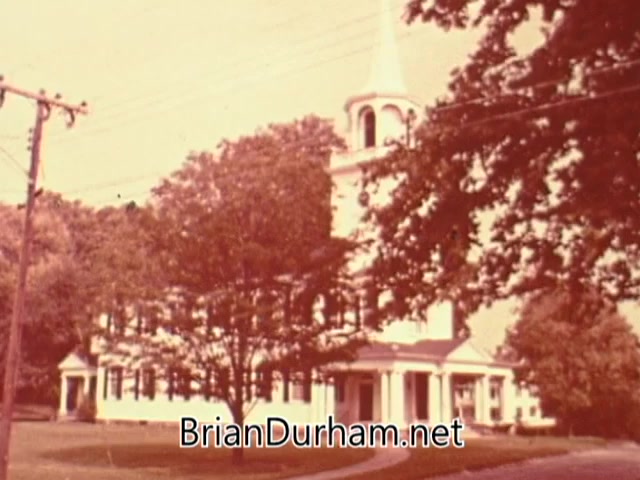 The still from the 'American Institute of Architects PSA' depicts a quaint, traditional building resembling a church, characterized by its tall steeple and white Colonial-style architecture. Surrounding the structure are lush green trees, suggesting a serene and inviting environment. The image reflects a sense of community and beauty, contrasting with the message of sprawl and lack of meaningful design found in modern housing developments. The overall atmosphere evokes nostalgia for a time when spaces fostered connection and aesthetic value.