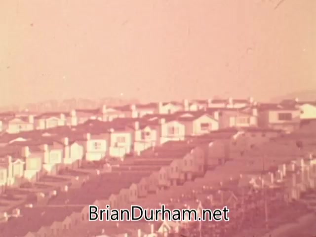 The still from the 1960s American Institute of Architects PSA depicts a vast expanse of suburban homes, characterized by uniformity and sprawl. The imagery emphasizes the contrast between the idealized American dream of community and beauty, and the reality of monotonous, disconnected housing developments. The muted colors suggest a lack of vibrancy and charm, reinforcing the message of lost potential in suburban design.