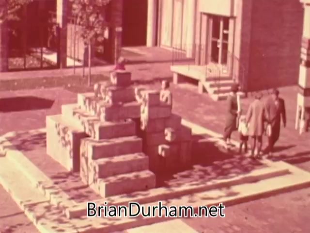 The still from the 1960s American Institute of Architects PSA depicts a small plaza featuring a sculptural installation resembling a stepped structure. A few figures are seen gathered around the installation, suggesting a social interaction space. The background includes modernist architecture, emphasizing the contrast between aesthetic community design and the sprawl criticized in the narrative, highlighting a vision for beautiful and functional urban living.