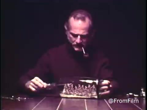 The still from 'C&P Telephone - 1969? Extension phones' features a man wearing a black sweater, focused on a transparent container filled with a series of interconnected parts, likely related to telephone equipment. He is seated at a table, holding a tool, with a cigarette in his mouth, creating a sense of concentration and craftsmanship. The overall ambiance suggests a DIY or repair context for telephone systems. The pre-1970 Bell System logo indicates its historical significance in telecommunications.   Follow @FromFilm for more archival insights!