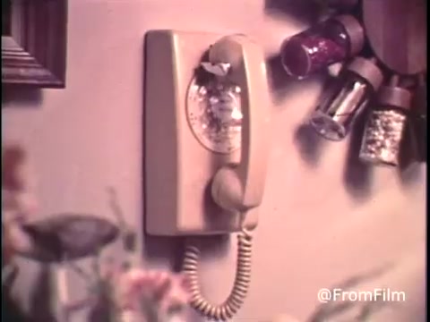 The still depicts a vintage telephone mounted on a wall, featuring a design characteristic of the late 1960s. The telephone is a cream color, with a distinct rotary dial and a curved handset. Nearby, various spice jars are visible, suggesting a domestic setting. The pre-1970 Bell System logo is present, highlighting its historical significance. This image reflects the era's home communication technology.   Follow us on Twitter @FromFilm for more vintage insights!