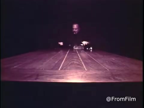 The still from 'C&P Telephone - 1969? Extension phones' features a dimly lit setting with a lone figure, likely a man, seated at a large wooden table. He appears to be engaged with a telephone on the table, suggesting a focus on communication technology. The pre-1970 Bell System logo indicates the piece's vintage, reflecting an era when extension phones were being introduced. The overall atmosphere feels serious and introspective, emphasizing the importance of telephonic connections during that time. Follow us on Twitter @FromFilm.