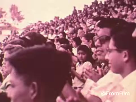 The still from the 'Project HOPE Public Service Announcement' features a large crowd gathered, likely for an event related to the organization. The audience appears engaged and diverse, showcasing a range of ages and expressions as they applaud. The overall atmosphere suggests a community event emphasizing health and hope, consistent with Project HOPE's mission to provide international healthcare. The color tone indicates a vintage style typical of the 1960s or 70s.