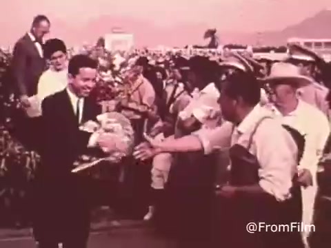 The still from the 'Project Hope Public Service Announcement - 60's/70's' depicts a moment of goodwill, likely showcasing the organization’s humanitarian efforts. A well-dressed man is seen shaking hands with a local individual in a casual outfit, possibly representing the community served by Project HOPE. The background features a gathering of people, suggesting a public event. This scene captures the spirit of collaboration and health care outreach central to Project HOPE's mission during that era. The color palette is likely warm, hinting at the nostalgic feel of the 60s and 70s.